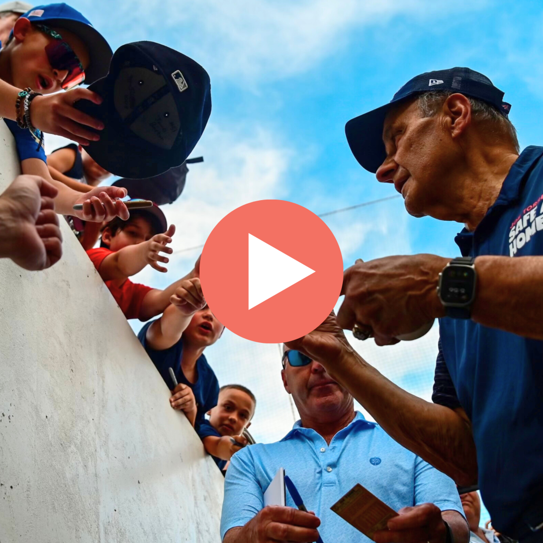 Website - baseball video play photo - Joe Torre Safe At Home Foundation Group of people leaning over a white barrier, reaching toward the camera at an outdoor event; a man in a blue shirt holds a booklet.